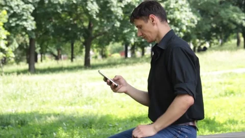 Guy sits in a park on a bench with a smartphone in his hand and looks at him Stock Footage 135238179