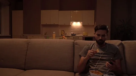 Guy sits on sofa and holds bpwl of chips. He is eating and listening to music Stock-Footage 99739112
