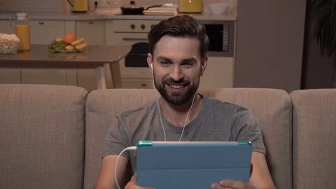 Guy sits on sofa and looks at tablet. He has headphones in ears. Bearded man is Stock Footage 99739146