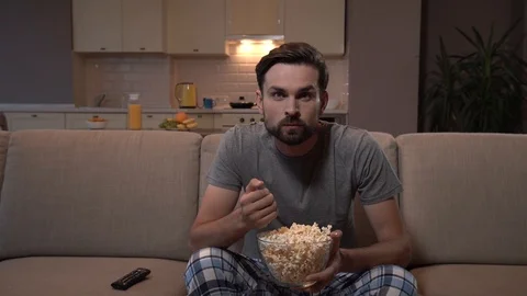 Guy sits on sofa and watches TV. He eats popcorn. Guy hs it in glass bowl. He is Stock Footage 99739506