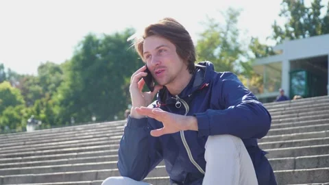 Guy sits on the steps and emotionally communicates on the phone in blue jacket Stock-Footage 163336228