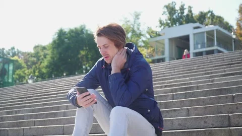 Guy sits on the steps and emotionally communicates on the phone in blue jacket 動画素材 163336282