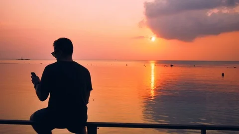 Guy is sitting on the beach and looking at the smartphone on the sunset Stock Footage 92227041