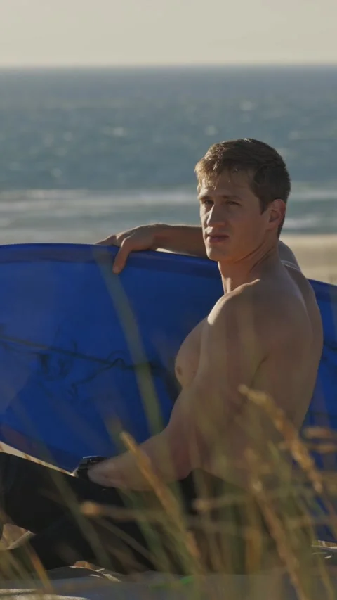 A guy sitting by the beach at early dawn holding a surfboard - looking in the Stock Footage 328670437