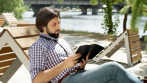 Guy is sitting on bench and reading a book. Then he got message on phone. Man is Stock-Footage 99739919