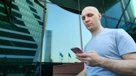 Guy sitting on bench near mirrored skyscraper and using mobile phone Stockbeeldmateriaal 201635921