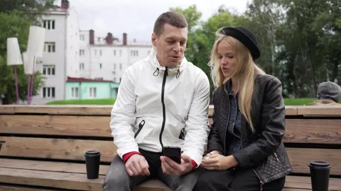 A guy sitting on a bench shows something on the phone to a girl. Video stock 263537620