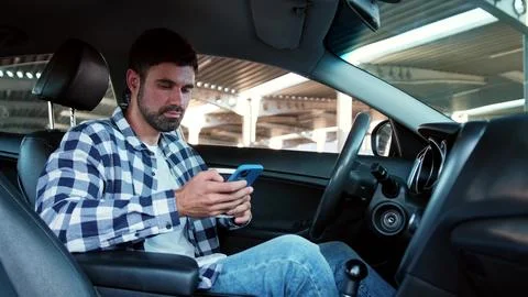 Guy sitting in the car while holding smartphone, typing, drinking coffee Stock Photos