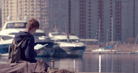 The guy is sitting in front of the sea and carefully reading a book Stock Footage 119613120