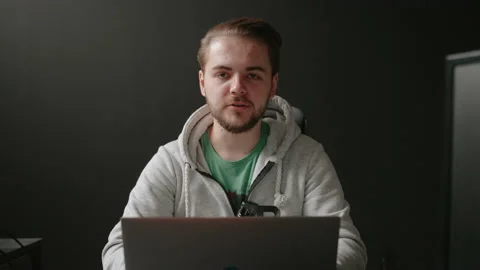 Guy sitting in isolated dark room behind laptop and talking to camera. Stock Footage 171713650