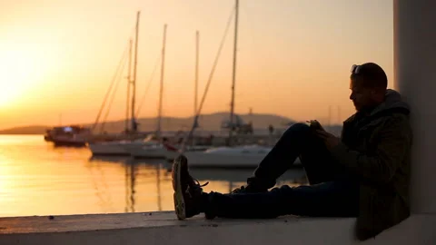 The guy is sitting on the parapet in the port At sunset Smoking Writing on Phone Video stock 150280480