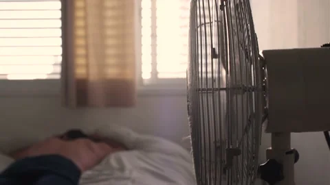 Guy sleeping in bed electric fan cooling the room and blowing wind on him. dusty Stock Footage 156935715