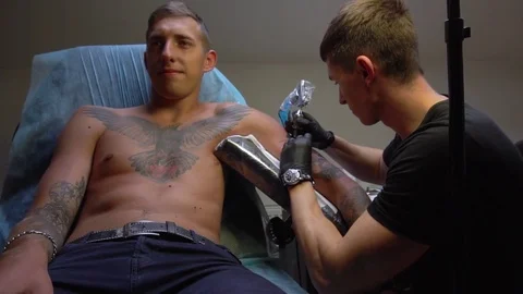 Guy smiles sitting in the chair while the master makes him a tattoo on his arm Stock-Footage 82840325