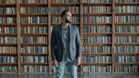 Guy Is Standing In The Library. Attractive Brunette With Beard From The Middle Stock Footage 139893556