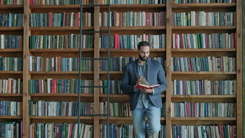 Guy Stands With His Back To The Shelves With Books And Reads Book. Stock Footage 140221056