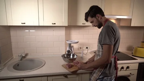 Guy stands in kitchen and cuts pieces of meat. Then he turns on meat grinder and Stock Footage 99739326
