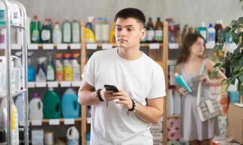 Guy in store scrolling through phone screen shopping list, woman shopping in Stock Photos