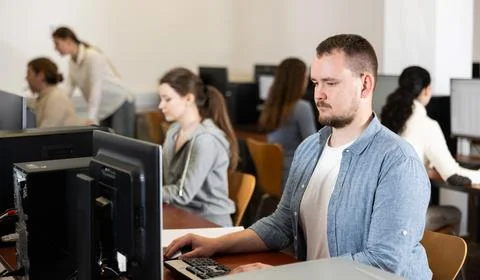 Guy student of computer courses looks at monitor screen, types on keyboard and Stock-Fotos