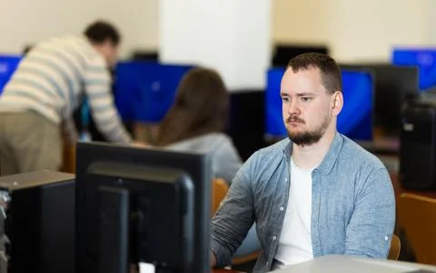 Guy student of computer courses looks at monitor screen, types on keyboard and Stock Photos