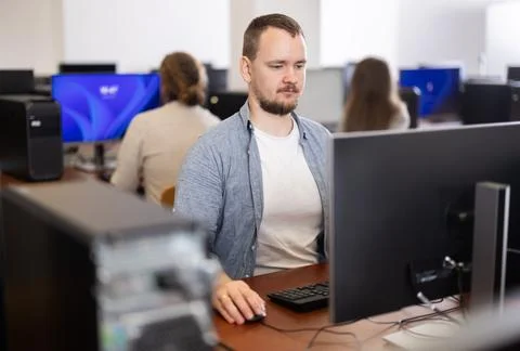 Guy student of computer courses looks at monitor screen, types on keyboard and Stock Photos