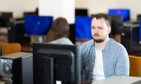 Guy student of computer courses looks at monitor screen, types on keyboard and Stock Photos