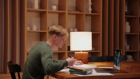 Guy studying at home, having a video call on a laptop, searches through textbook Stock-Footage 164380569