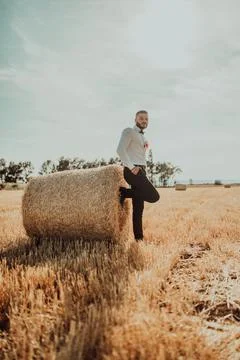 A guy in a suit posing in front of a big bale of hay in a field at sunset Stock Photos
