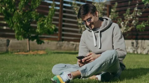 Guy in sunglasses sitting on the grass with a mobile phone looking at the camera Stock Footage 247966931