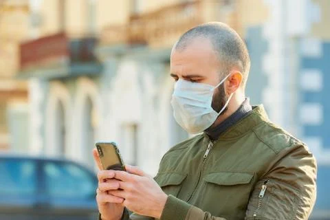 A guy with a surgical mask on the face using a smartphone on a street. Stock Photos