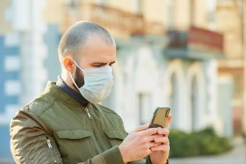 A guy with a surgical mask on the face using a smartphone on a street. Stock Photos
