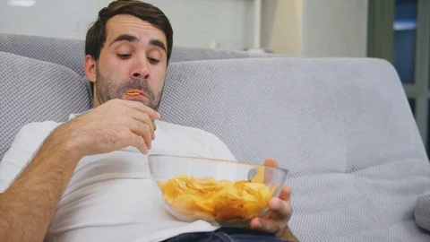 The guy in the T-shirt sits on the couch, eats chips and watches the TV. While Video stock 104011802