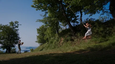 Guy throws a hat on the head of a girl swinging on a swing in a nature park 库存影片 127543780