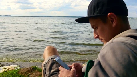 The guy uses the phone while sitting on a chair by the lake. Stock Footage 77759817