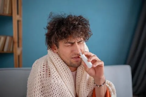 Guy using nose spray. Photo of sick arabic man covered plaid at home. Medical Stock Photos