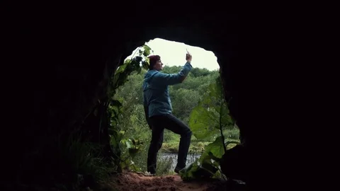Guy using smartphone standing near exit from cave. Deep forest at background. Stock Footage 80053844