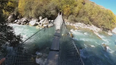 A guy walks on a suspension bridge passing through a mountain river Stock Footage 183055029