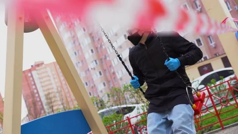 Guy wearing mask rides red swing on closed children playground during pandemic Video stock 131477402