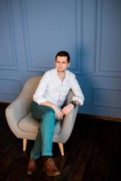 A guy, wearing a white shirt with patterns and green pants, sitting in armchair 库存照片
