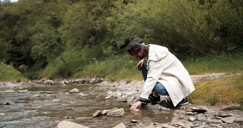 A guy in a white hiking jacket draws water into a thermos from a mountain river Stock Footage 247051024