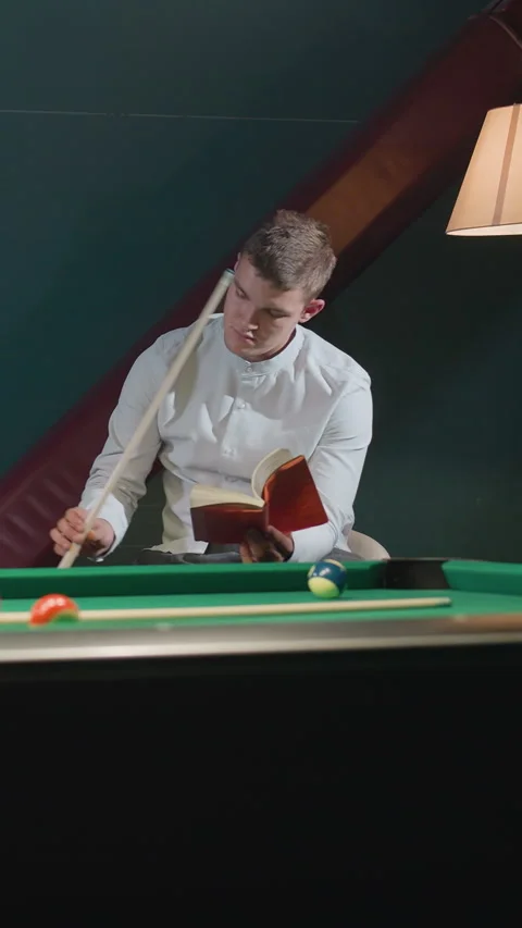 Guy in white shirt reading red book while holding cue stick near green pool Stock Footage 319808182