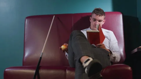 Guy in White Shirt Sitting on Red Sofa Reading Book Holding Billiard Ball Video stock 302012802