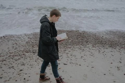 A guy in a winter jacket walks along the beach with a book in his hands Stock Photos