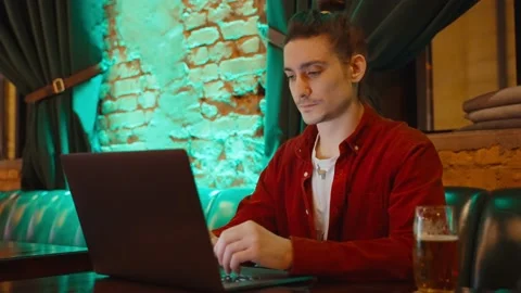 A guy working on a laptop sitting at a table in a cafe and drinking Stock Footage 231889132