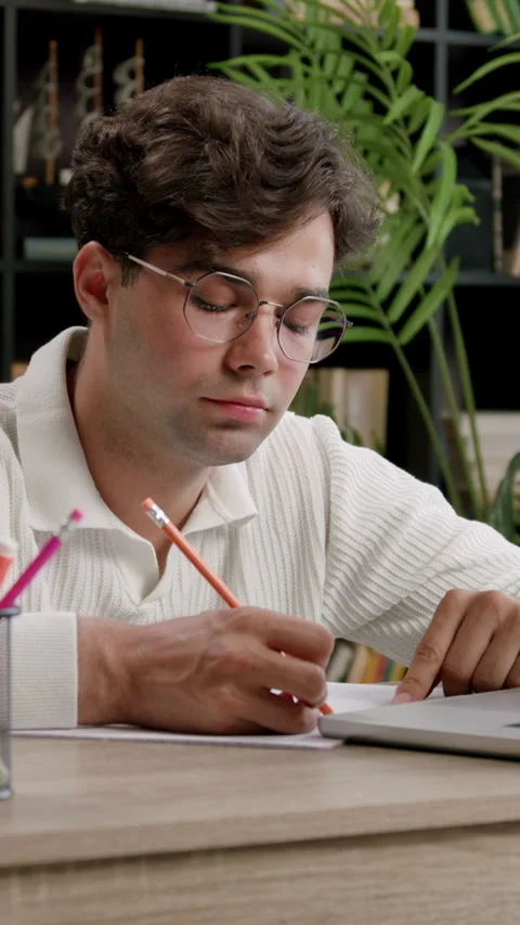 Guy writes with pencil on paper in front of open laptop Stock Footage 313930227