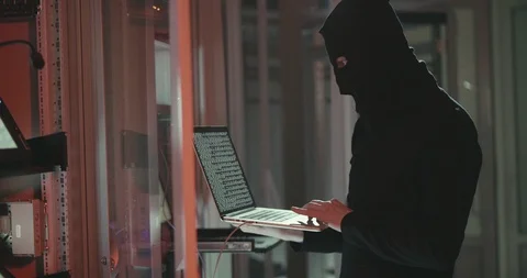 Hacker in black mask typing code in program using laptop in data center Stock Footage 107902985