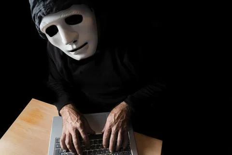 Hacker dressed in black with mask and hood typing on a computer while looking Stock Photos