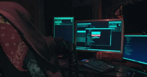 Hacker in a hood sits in front of computer screens and hacks databases Stock Photos