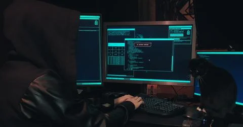 Hacker in a hood sits in front of computer screens and hacks databases Stock Photos