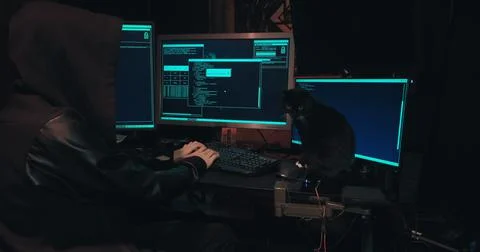Hacker in a hood sits in front of computer screens and hacks databases Stock Photos
