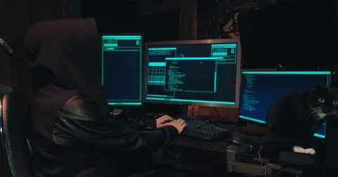 Hacker in a hood sits in front of computer screens and hacks databases Stock Photos
